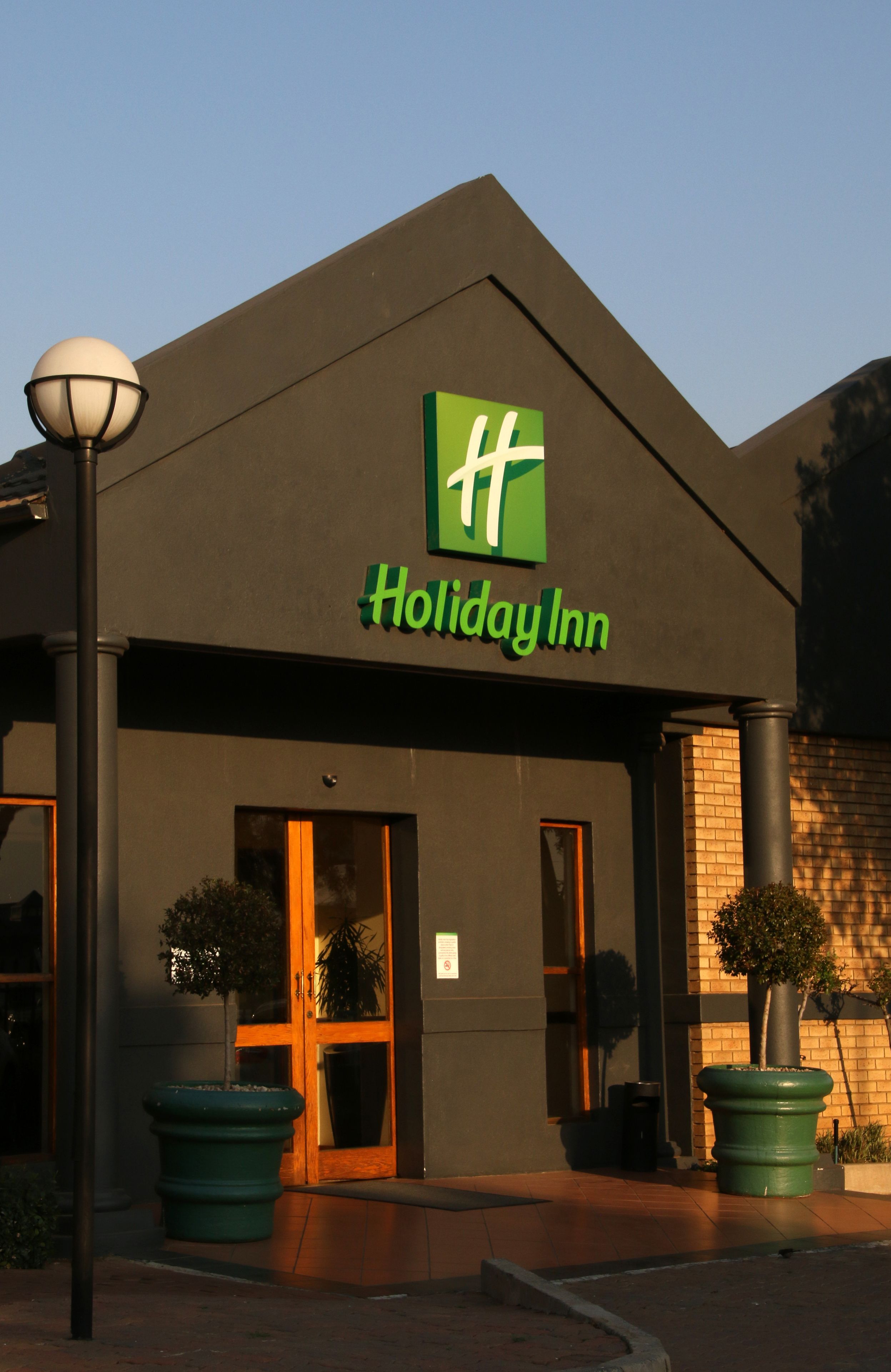 A Holiday Inn hotel entrance with a prominent green logo, large windows, and potted plants.