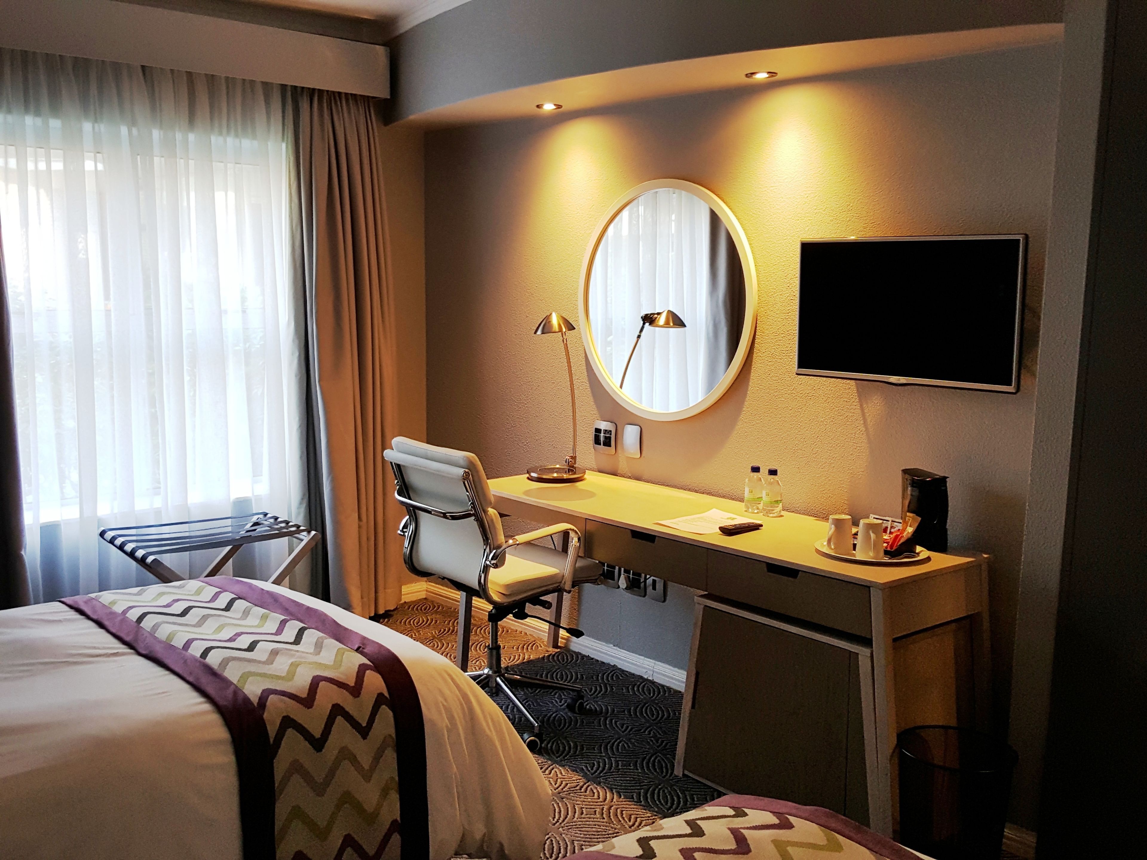 A hotel room with a bed, a desk, a chair, a round mirror, a television, and a window with curtains.