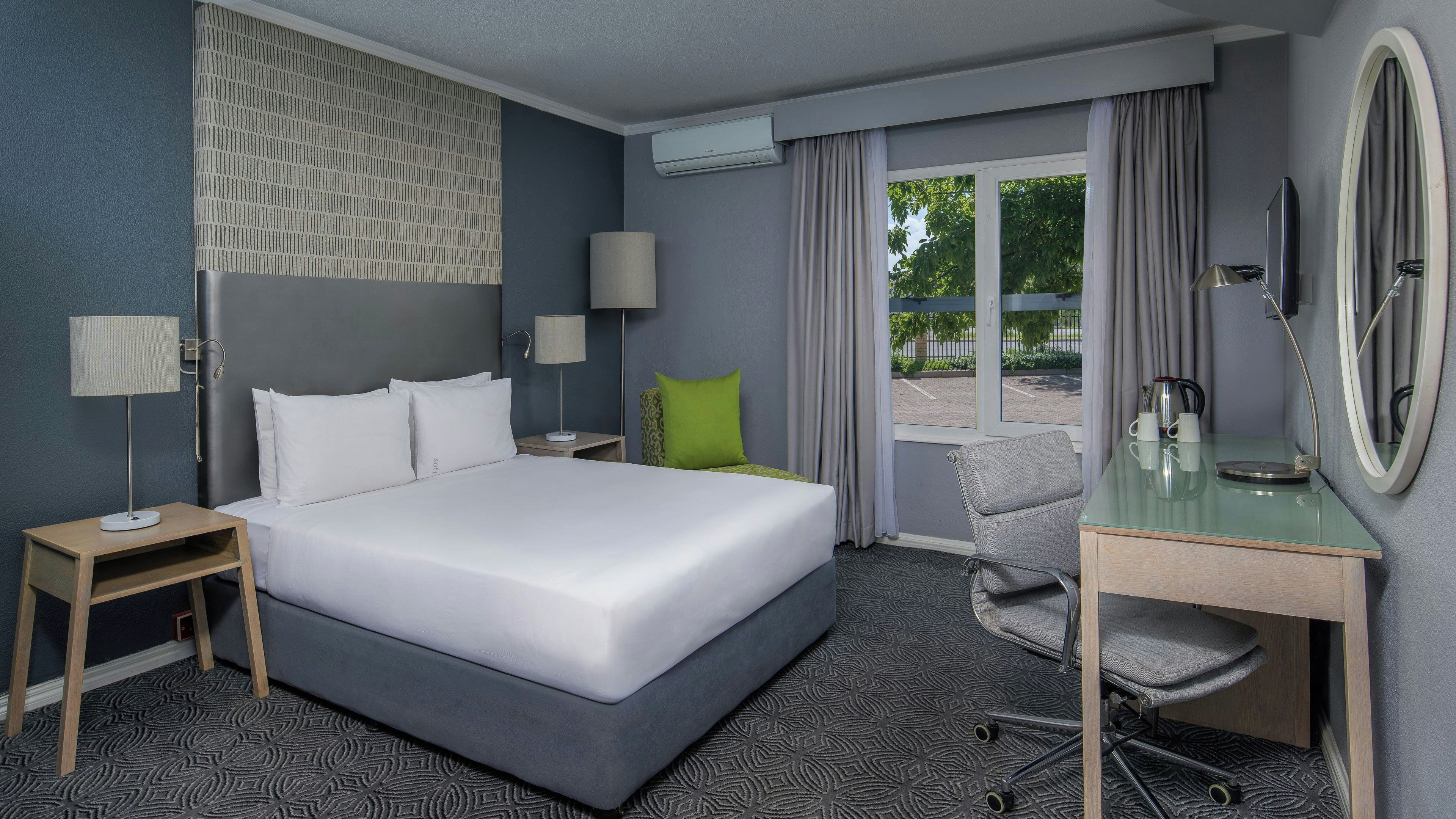 A hotel room with a bed, a desk, a chair, a lamp, an air conditioning unit, and a window with curtains.