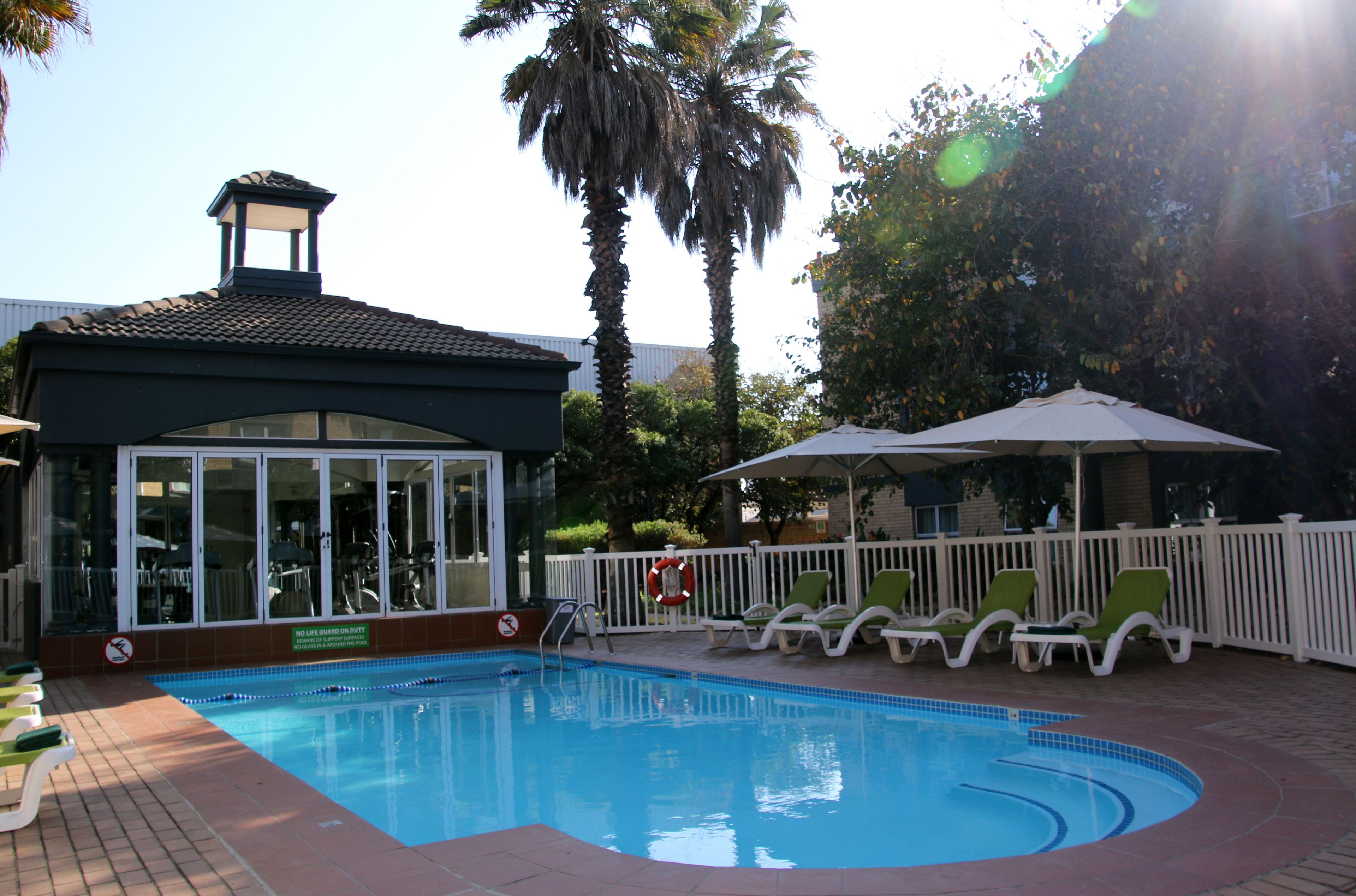 A swimming pool with lounge chairs and a covered area, surrounded by palm trees and a white fence.