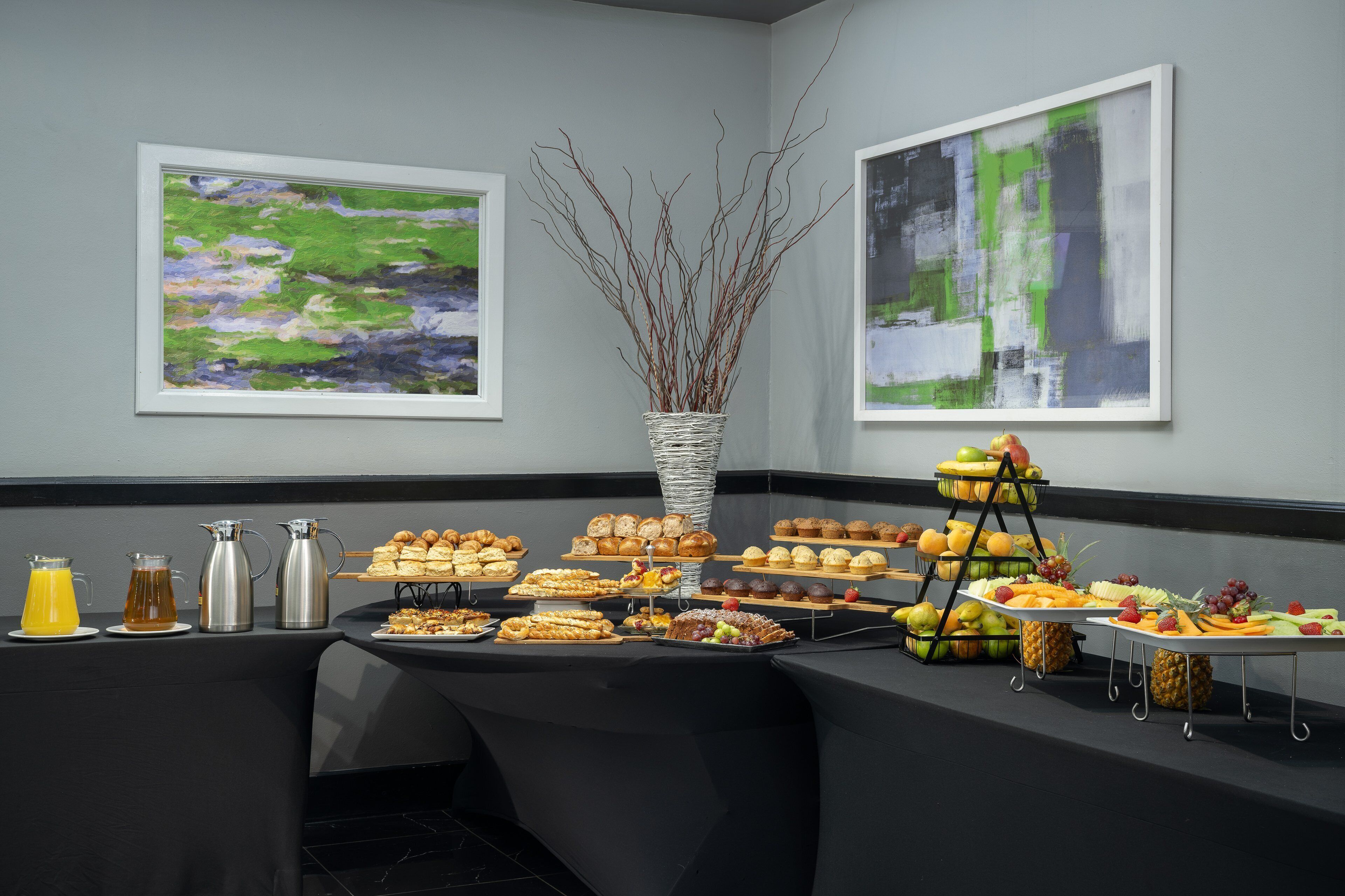 Une table de buffet garnie de divers plats et boissons, des œuvres d’art encadrées accrochées aux murs et un vase décoratif orné de branches.