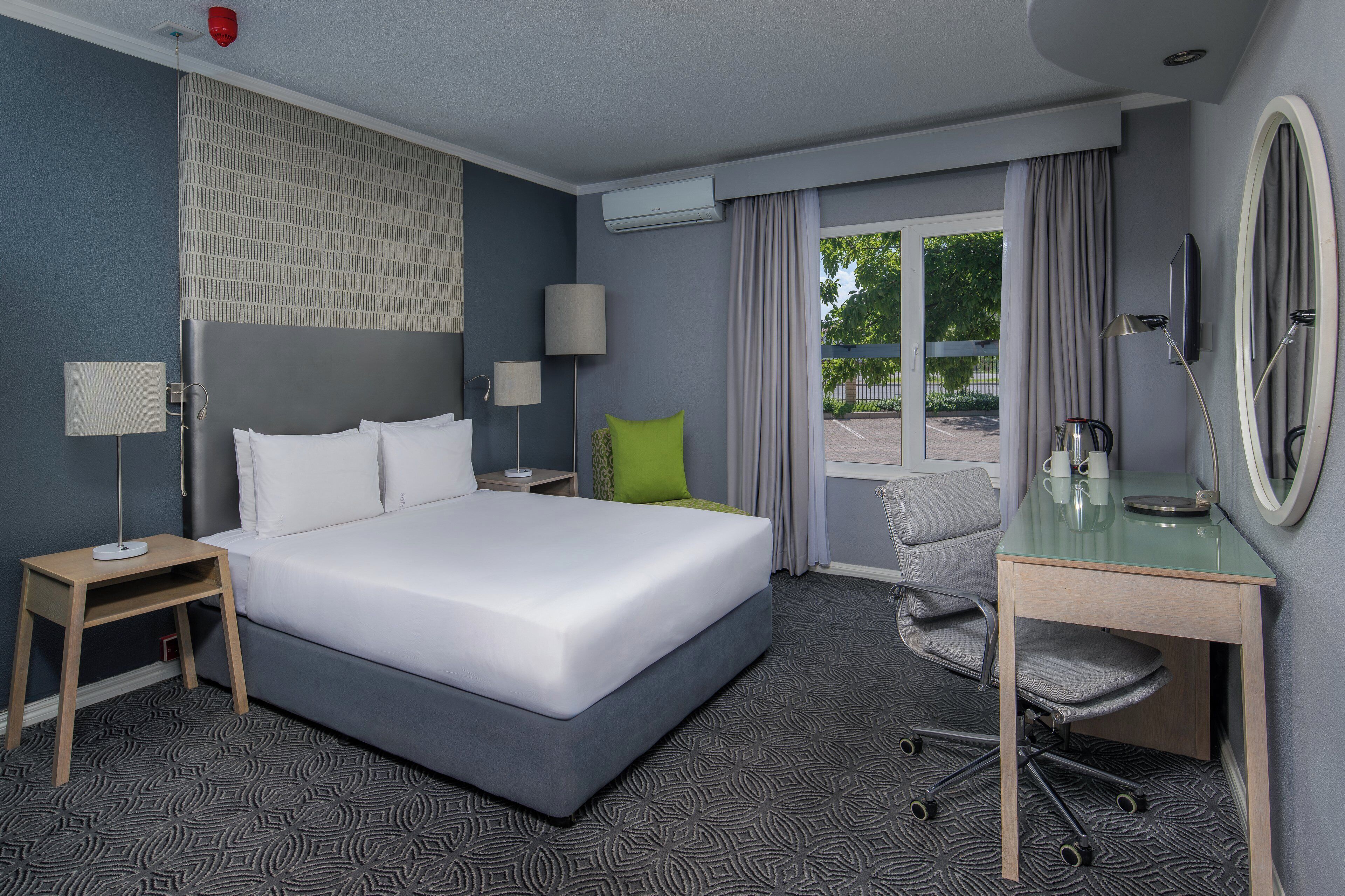 A hotel room with a large bed, a desk with a mirror, two bedside tables, a chair, and a view of the outdoors.
