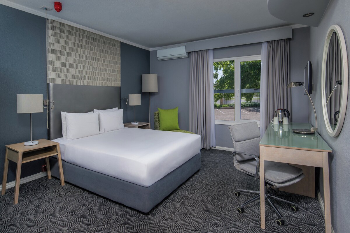 A hotel room with a large bed, a desk with a mirror, two bedside tables, a chair, and a view of the outdoors.
