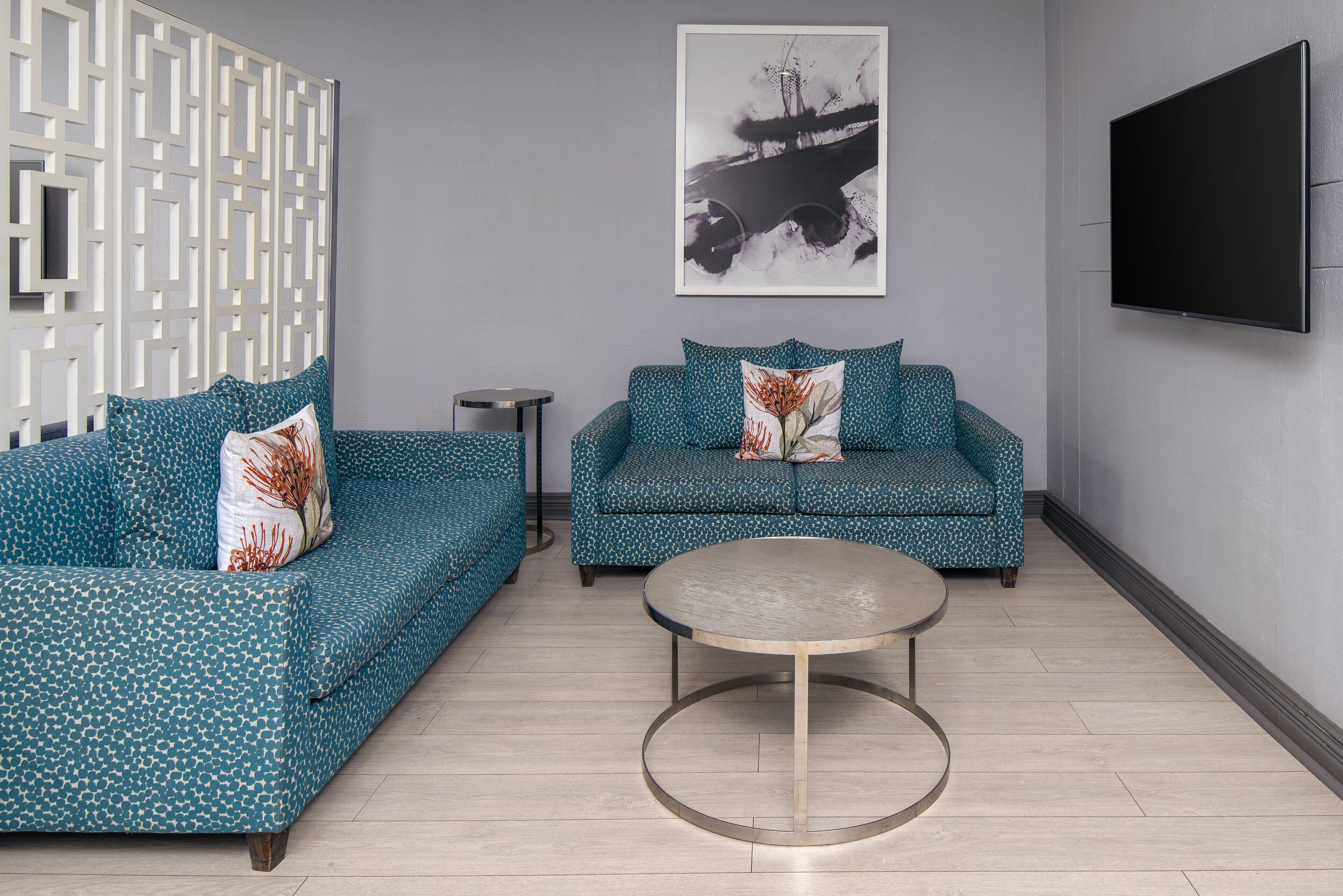 A modern living room with two teal patterned sofas, a round coffee table, a framed abstract artwork, and a flat-screen TV mounted on the wall.