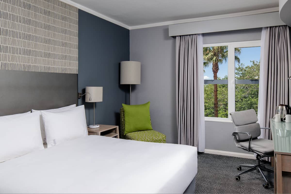 A hotel room with a large bed, a desk with a chair, a lamp, a window with curtains, and a view of trees.