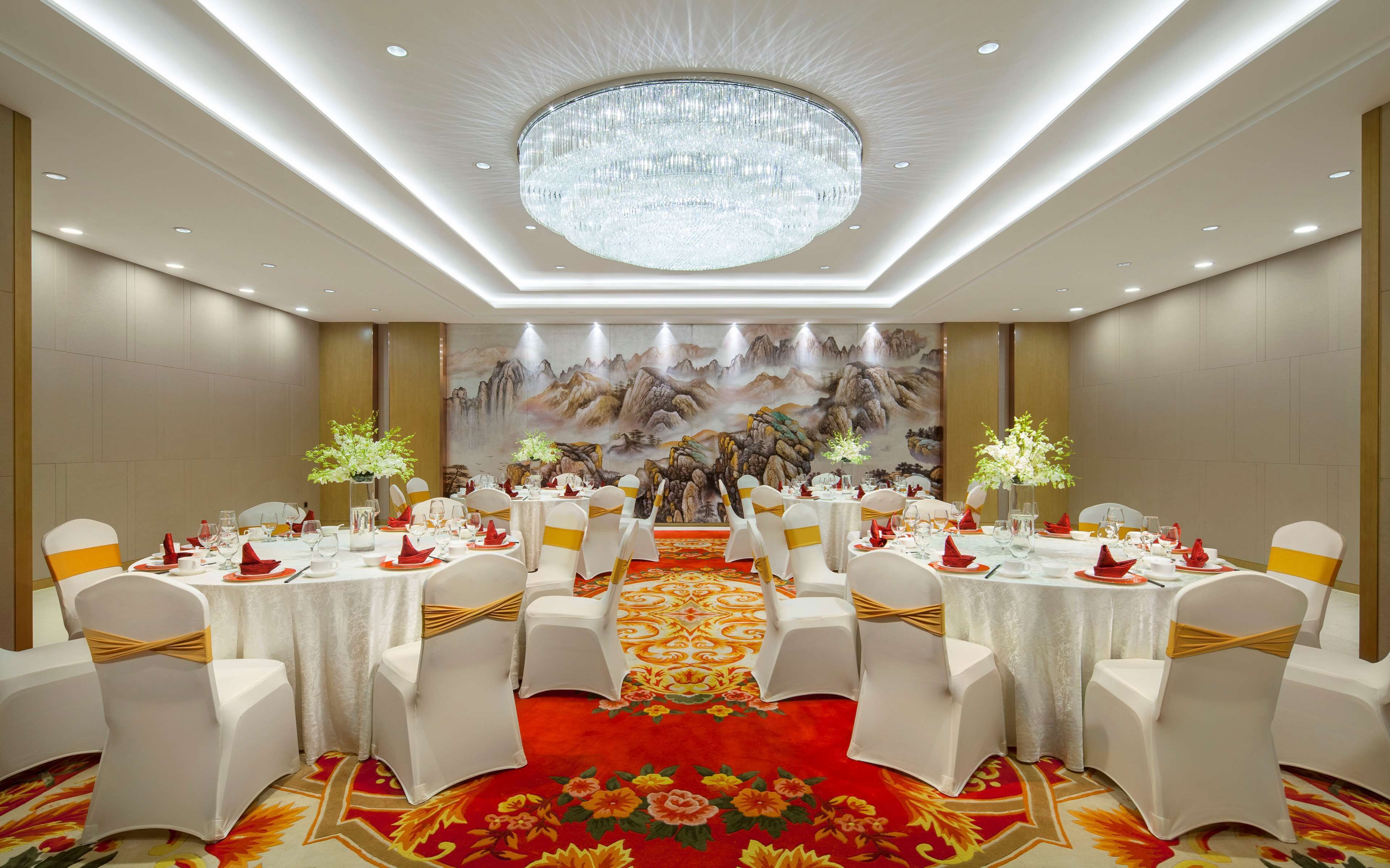 A banquet hall with round tables, white chairs with gold sashes, a red carpet, and a large chandelier.