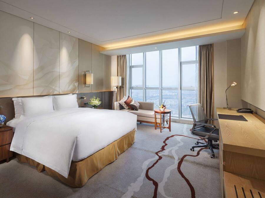 Una habitación de hotel moderna con una cama grande, un escritorio, una silla, un televisor y vistas a la ciudad a través de amplias ventanas.