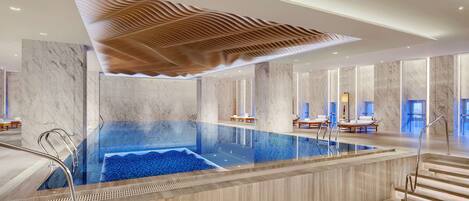 A modern indoor swimming pool with a wooden ceiling design, marble walls, and a seating area with loungers.