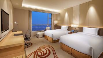 A hotel room with two beds, a desk, a chair, a TV, and a large window with a city view.