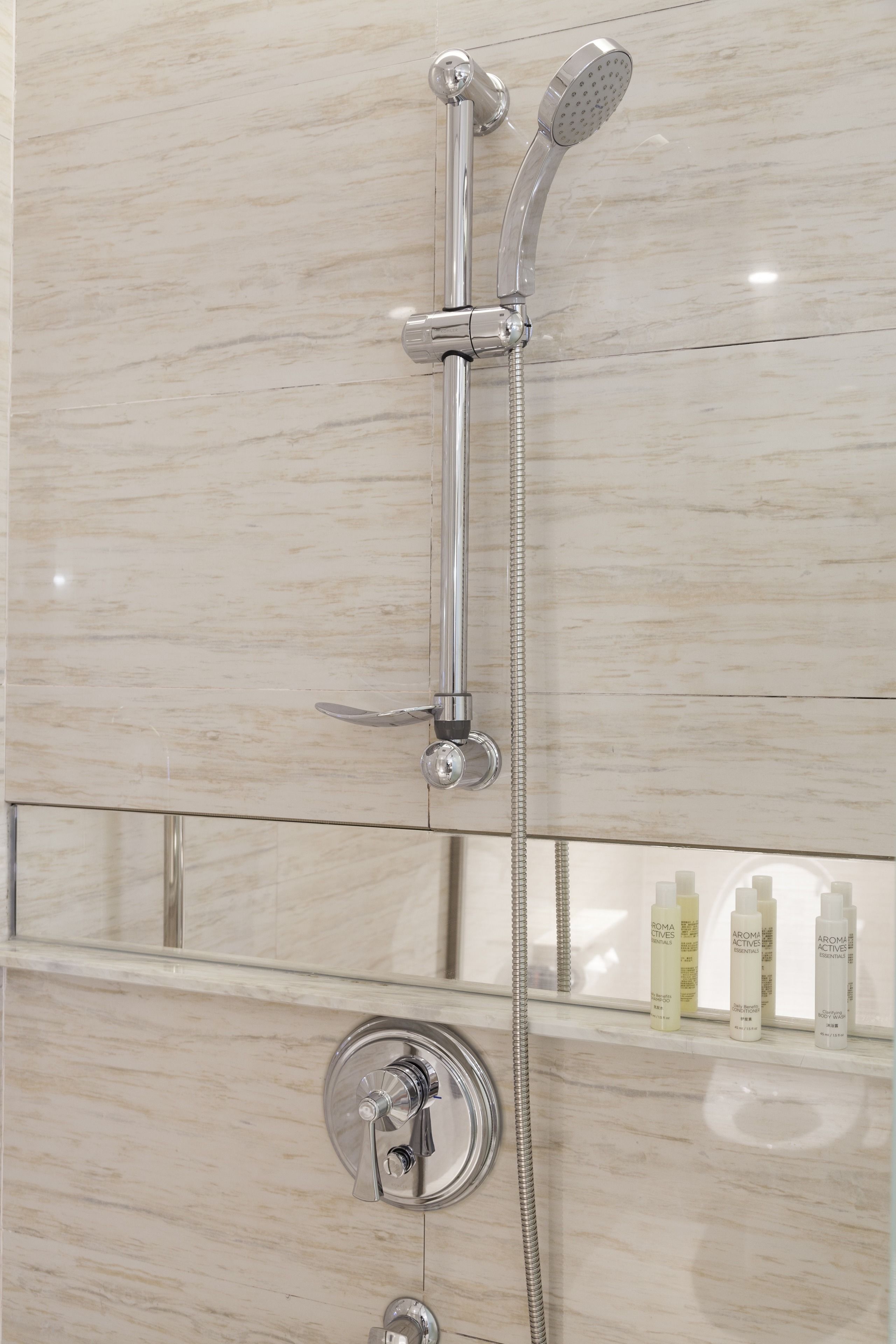 A shower with a handheld and a fixed showerhead, a control knob, and a glass shelf with toiletries.
