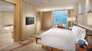 Una habitación de hotel moderna con una cama grande, un televisor montado en la pared, vistas a la ciudad y una pequeña mesa con un jarrón de flores.