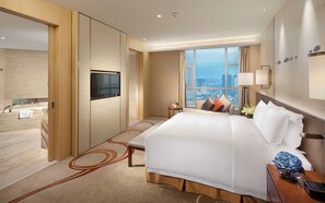 Una habitación de hotel moderna con una cama grande, un televisor montado en la pared, vistas a la ciudad y una pequeña mesa con un jarrón de flores.