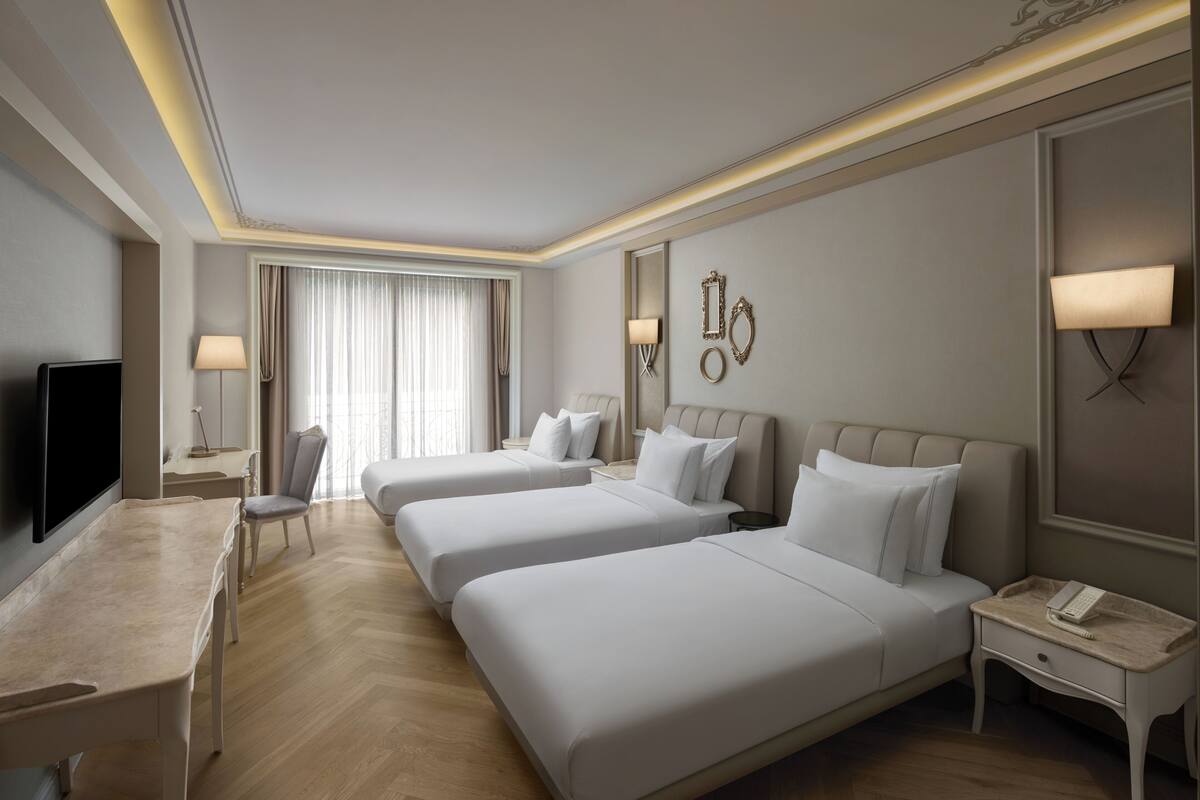 A hotel room with two beds, a desk, a chair, a TV, and a bedside table with a lamp.