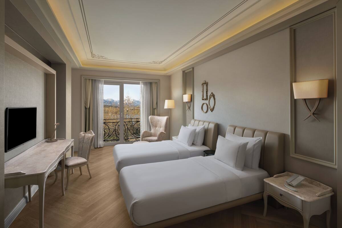 A hotel room with two beds, a desk, a chair, a nightstand with a phone, and a balcony with a view.