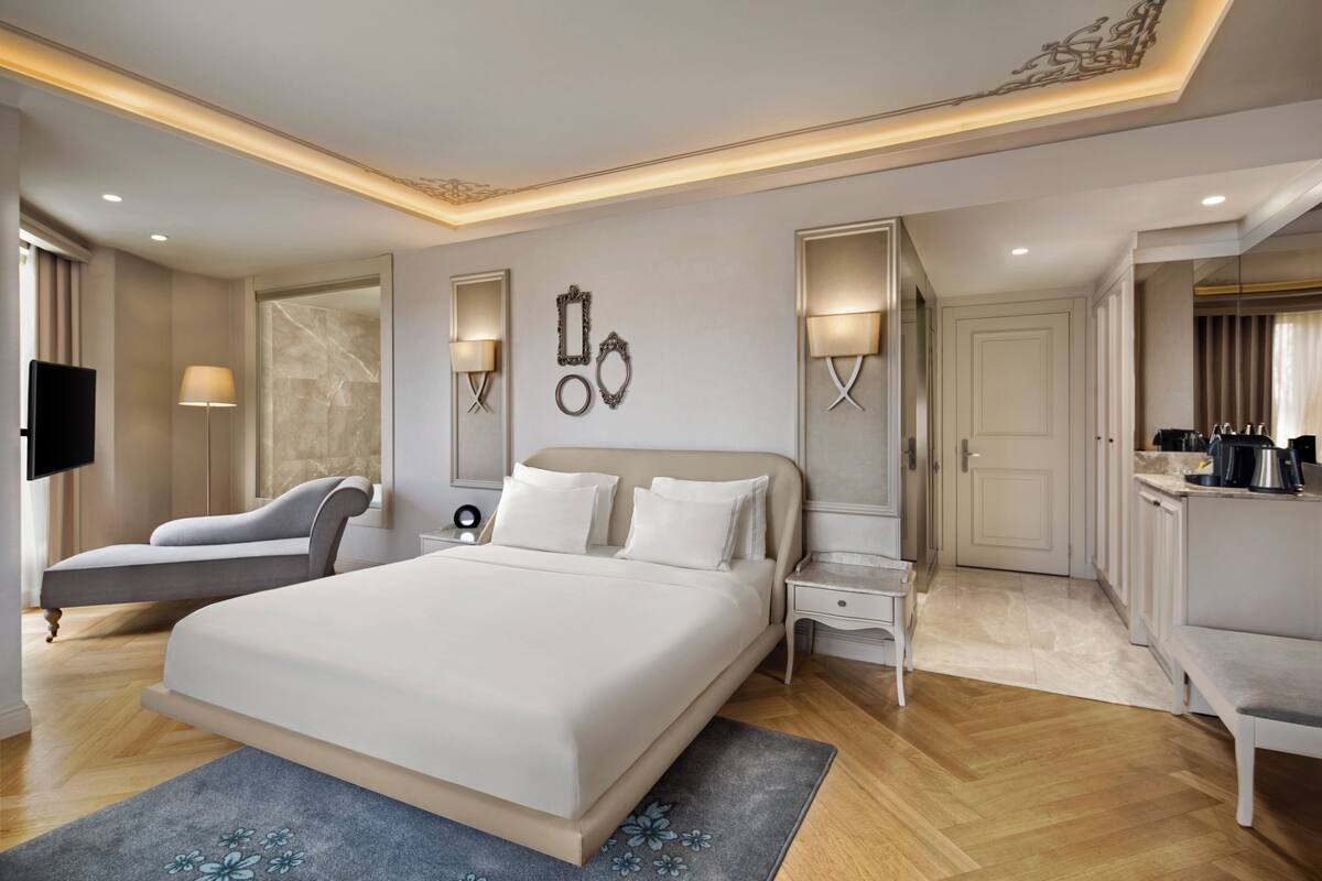 A spacious bedroom with a large bed, bedside tables, a flat-screen TV, and a seating area with a sofa and armchair.