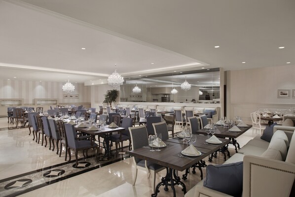 Un comedor espacioso con varias mesas y sillas, candelabros elegantes y una cocina moderna al fondo.