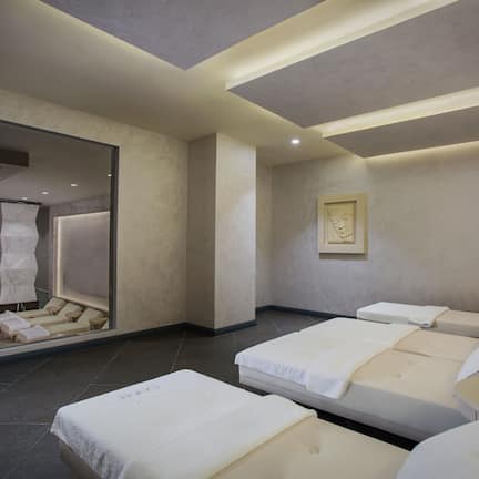A modern spa room with a large mirror, two massage beds, a small table with a glass and apple, and a framed artwork on the wall.