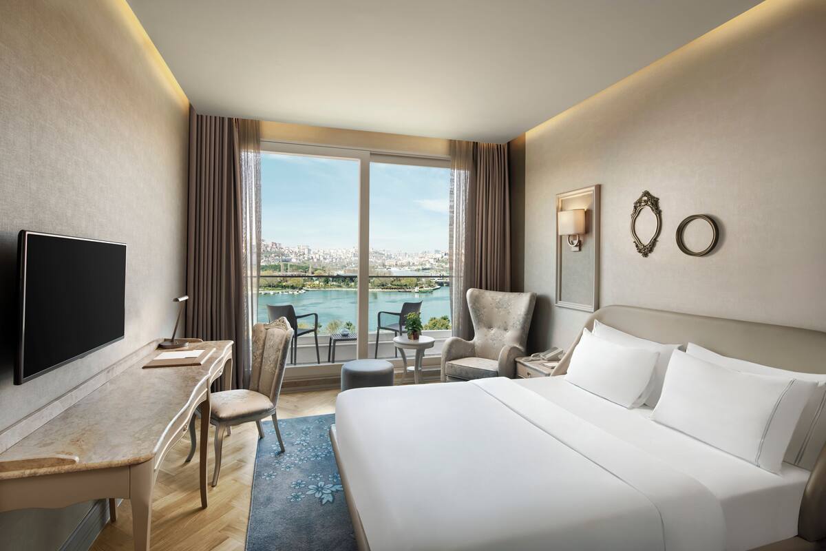 A hotel room with a bed, desk, TV, chair, and a view of a body of water.