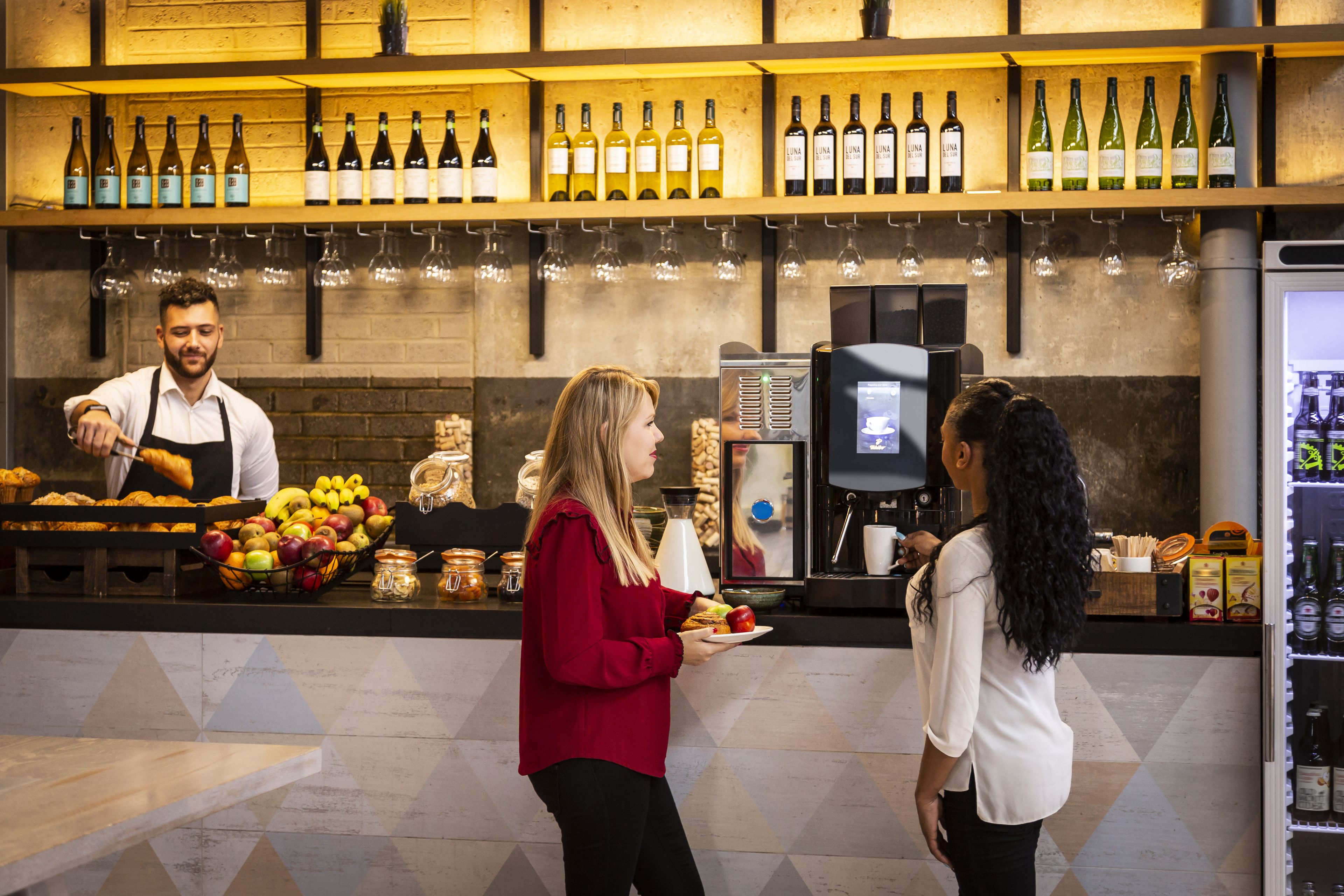 Un espace bar avec des étagères remplies de bouteilles de vin, un appareil à café et un présentoir de fruits.