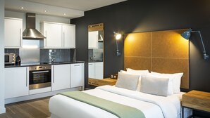 Een moderne hotelkamer met een groot bed, een houten hoofdbord, een nachtkastje, een spiegel en een kitchenette met een kookplaat, oven en magnetron.