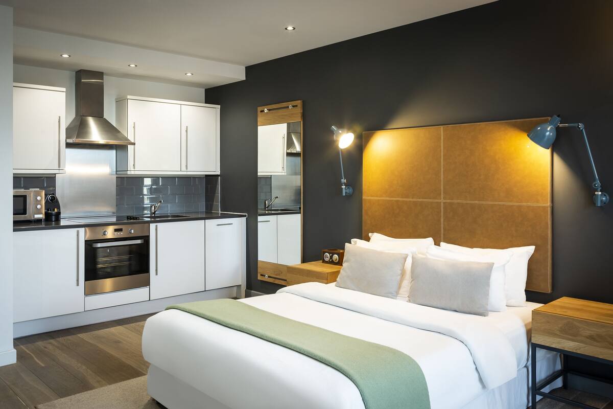 Une chambre d’hôtel moderne avec un grand lit, une tête de lit en bois, une table de chevet, une commode, un miroir et un coin cuisine équipé d’une cuisinière, d’un four et d’un four à micro-ondes.