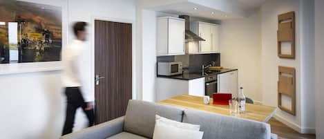 Un salon moderne avec un canapé gris, une table à manger en bois et un coin cuisine doté de meubles blancs et d’une hotte de cuisine en acier inoxydable.