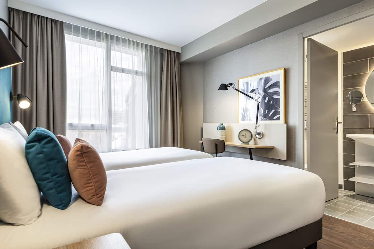Une chambre d’hôtel avec deux lits, un bureau avec une lampe, une horloge et un tableau encadré au mur.