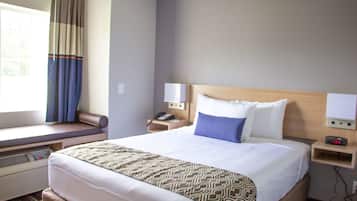 A hotel room with a large bed, a bedside table with a lamp, a window with curtains, and a wall-mounted heater.
