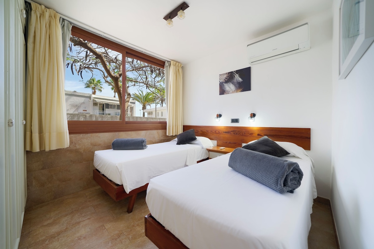A hotel room with two beds, a window with curtains, an air conditioning unit, and a view of palm trees and buildings outside.