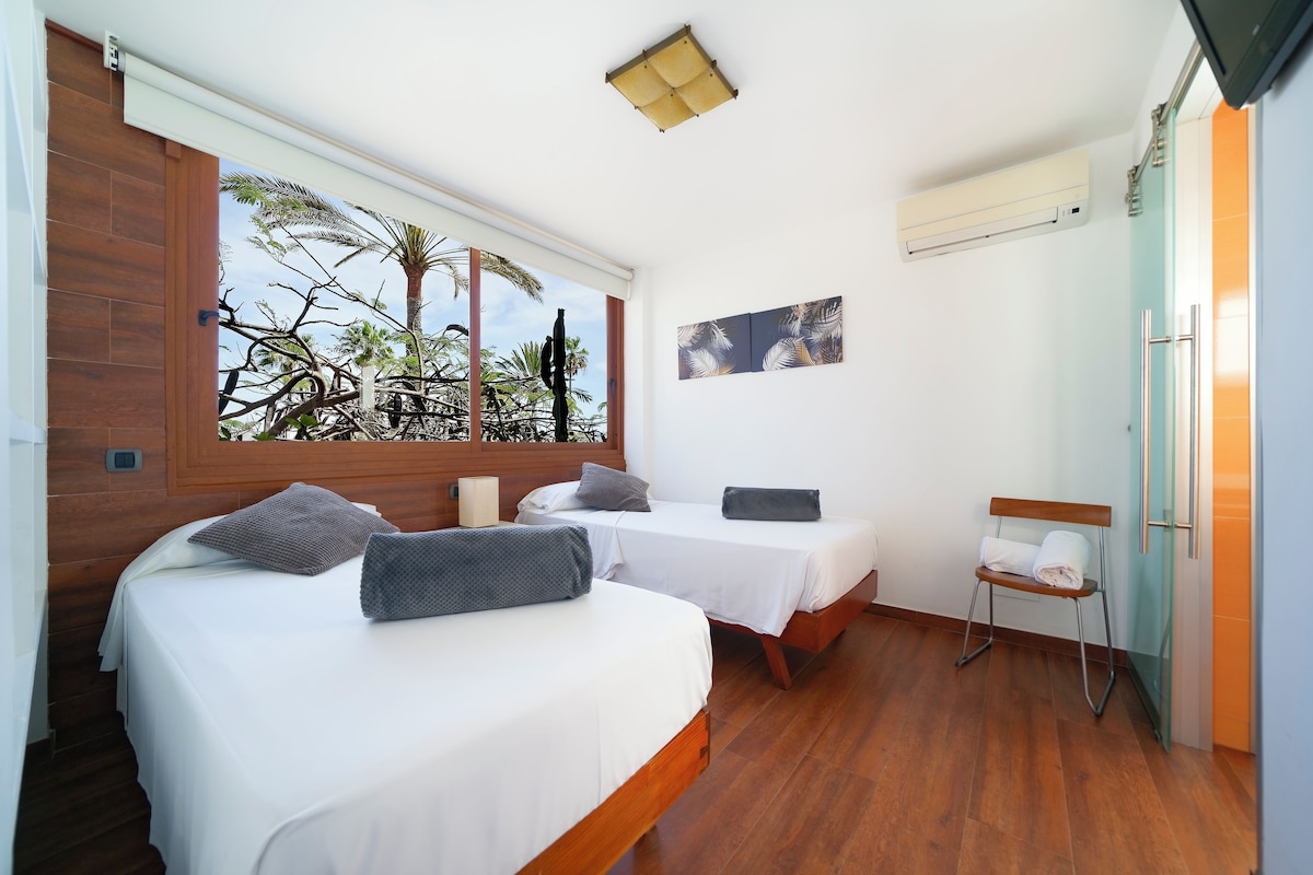 A room with two beds, a window with a view of palm trees, a small table, and an air conditioning unit.