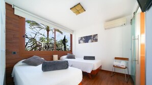 Una habitación con dos camas, una ventana que da a palmeras, una mesita y un banco.