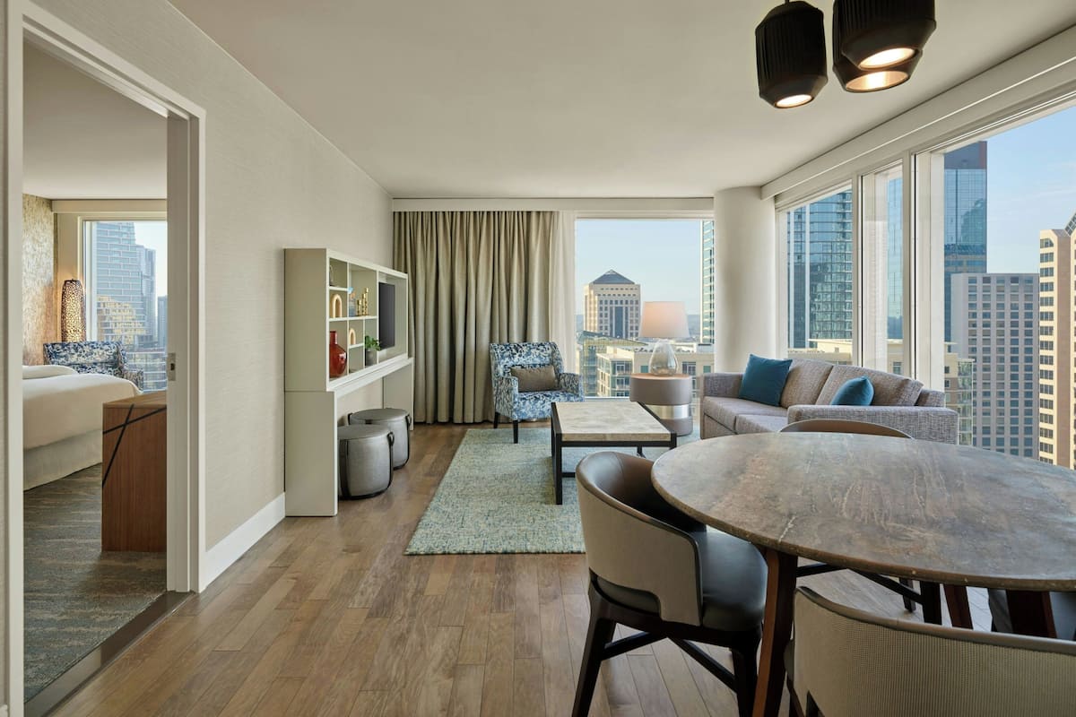 A modern living room with a sofa, armchairs, a dining table, and a bookshelf. Large windows offer a city view.