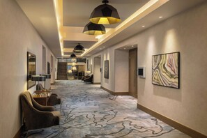 A hotel corridor with a patterned carpet, modern pendant lighting, and a large abstract painting on the wall.