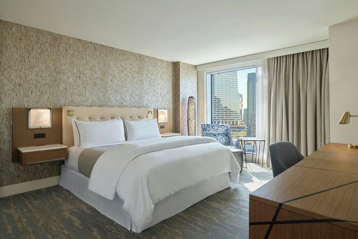 A hotel room with a large bed, bedside tables with lamps, a desk, and a view of the city through large windows.
