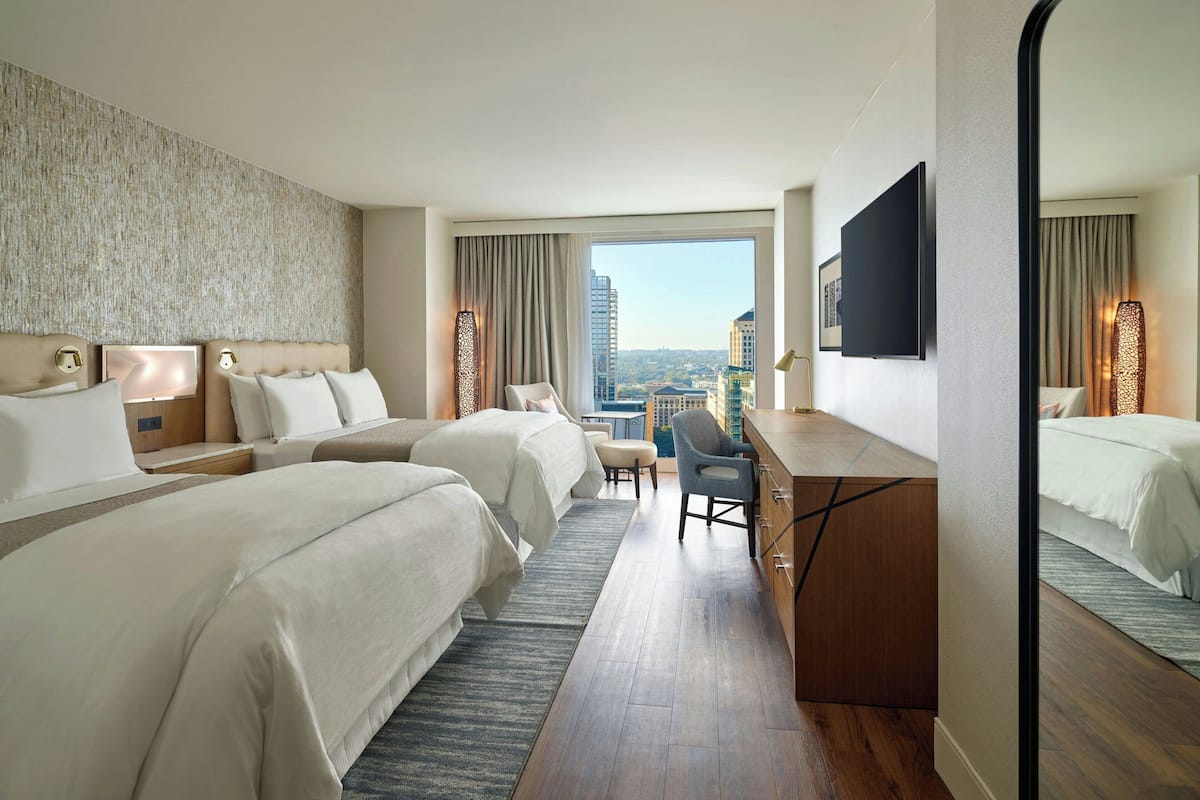 A hotel room with two beds, a desk, a chair, and a large window offering a city view.