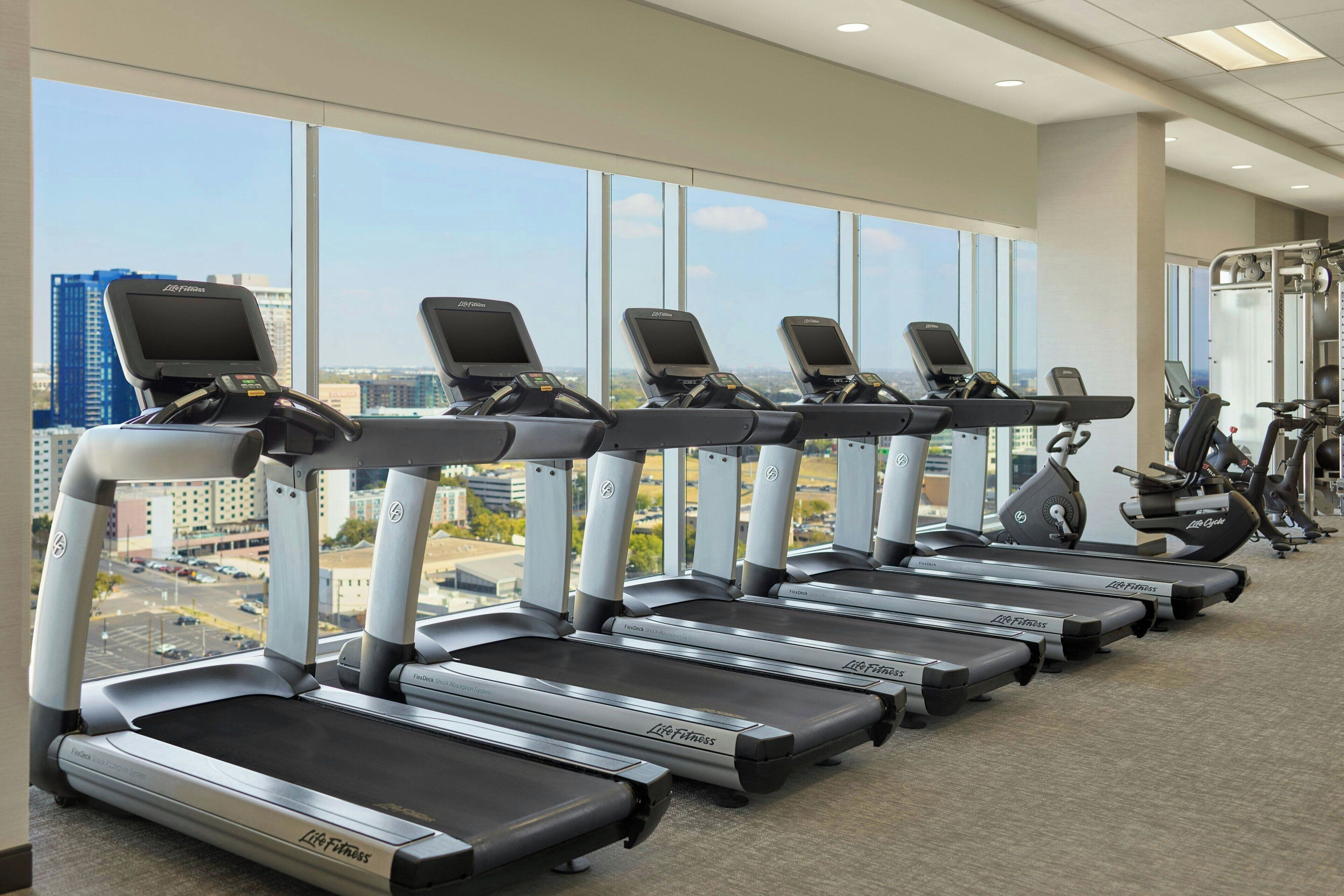 A row of treadmills with digital screens, a weight machine, and a large window with a city view.