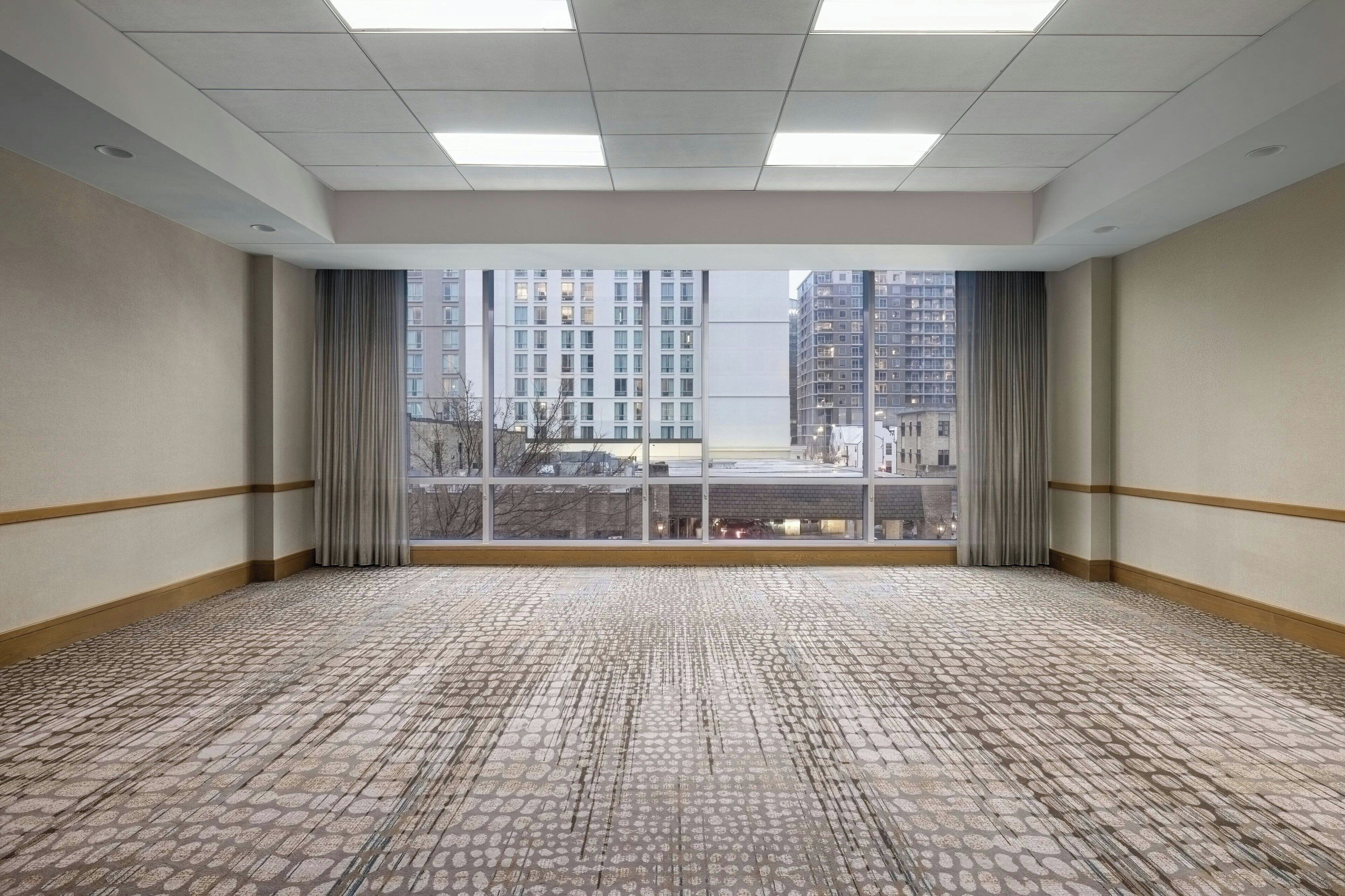 A large, empty room with patterned carpet, a view of tall buildings through a large window, and a ceiling with recessed lighting.