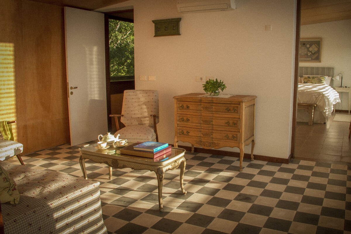 A room with a checkered floor, a wooden dresser, a chair, a table with books and a teapot, and a bed in the background.