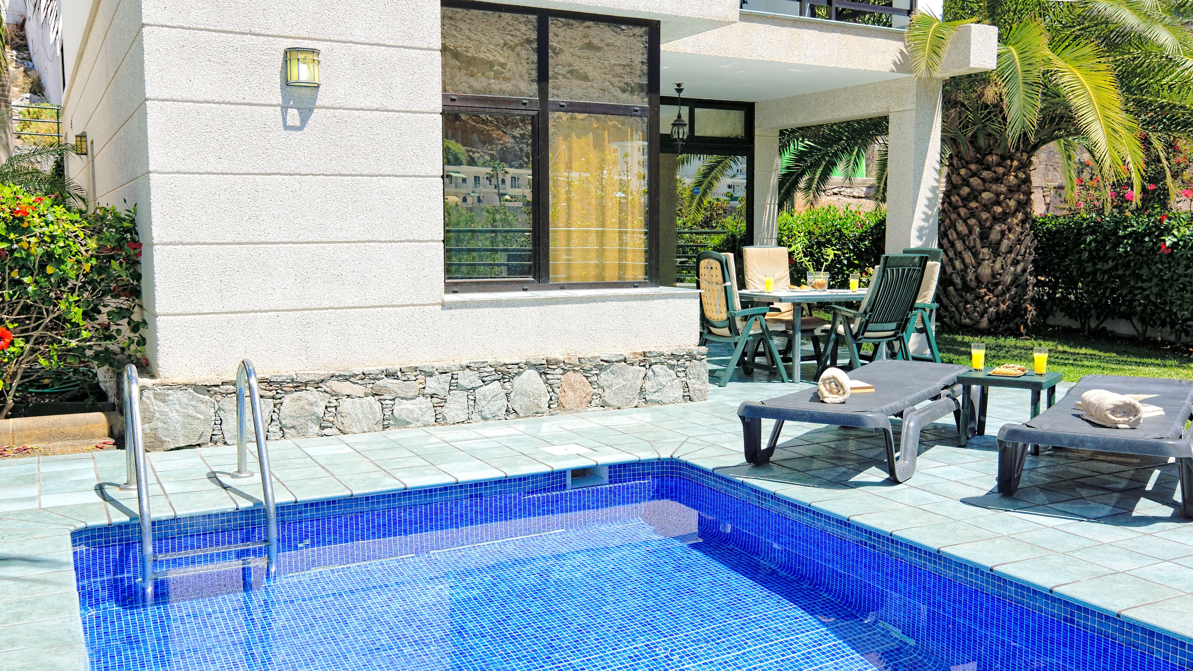 Ein Swimmingpool mit Liegestühlen und einem Tisch, umgeben von einer Steinmauer und Bepflanzung.