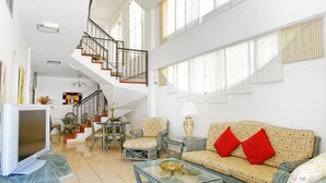 Un salon spacieux avec un plafond haut, un escalier, un canapé avec des coussins rouges et un téléviseur.