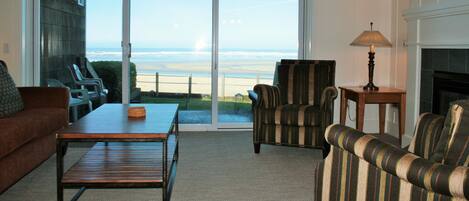 A living room with a large sliding glass door leading to a balcony with a view of the ocean. There is a fireplace, a lamp, and two striped armchairs.