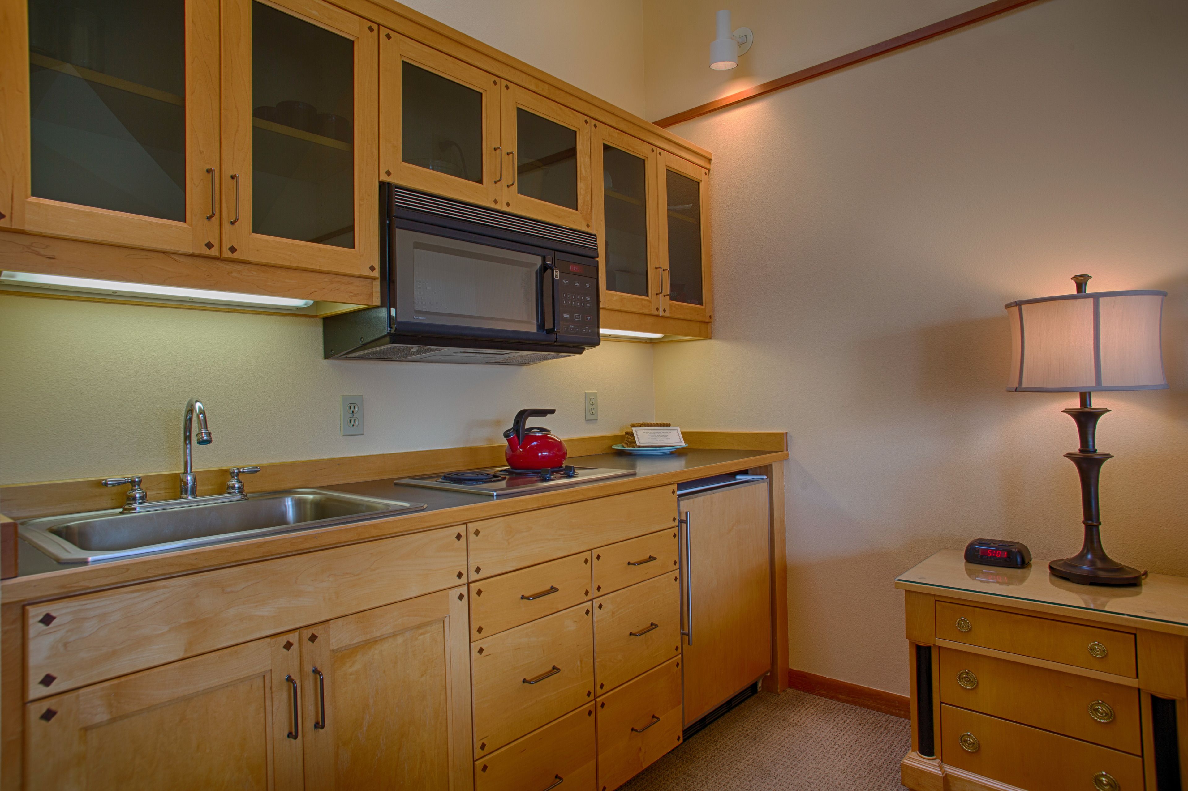 A kitchen with wooden cabinets, a microwave, a sink, a red kettle, and a lamp on a side table.
