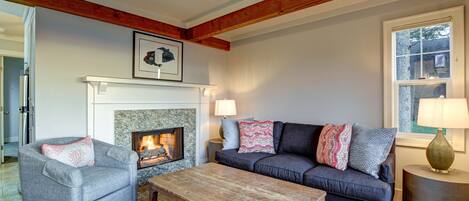 A cozy living room with a fireplace, a gray sofa with patterned cushions, a wooden coffee table, and a lamp on a side table.