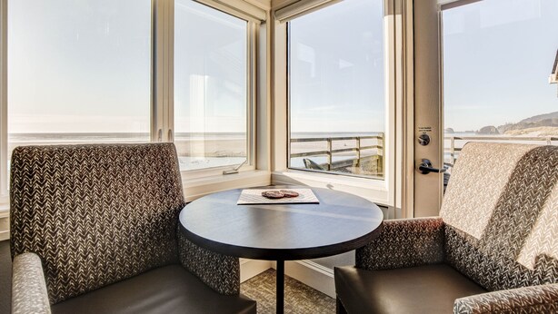 A room with a round table and two patterned armchairs, large windows with a view of the beach and ocean, and a ceiling light fixture.
