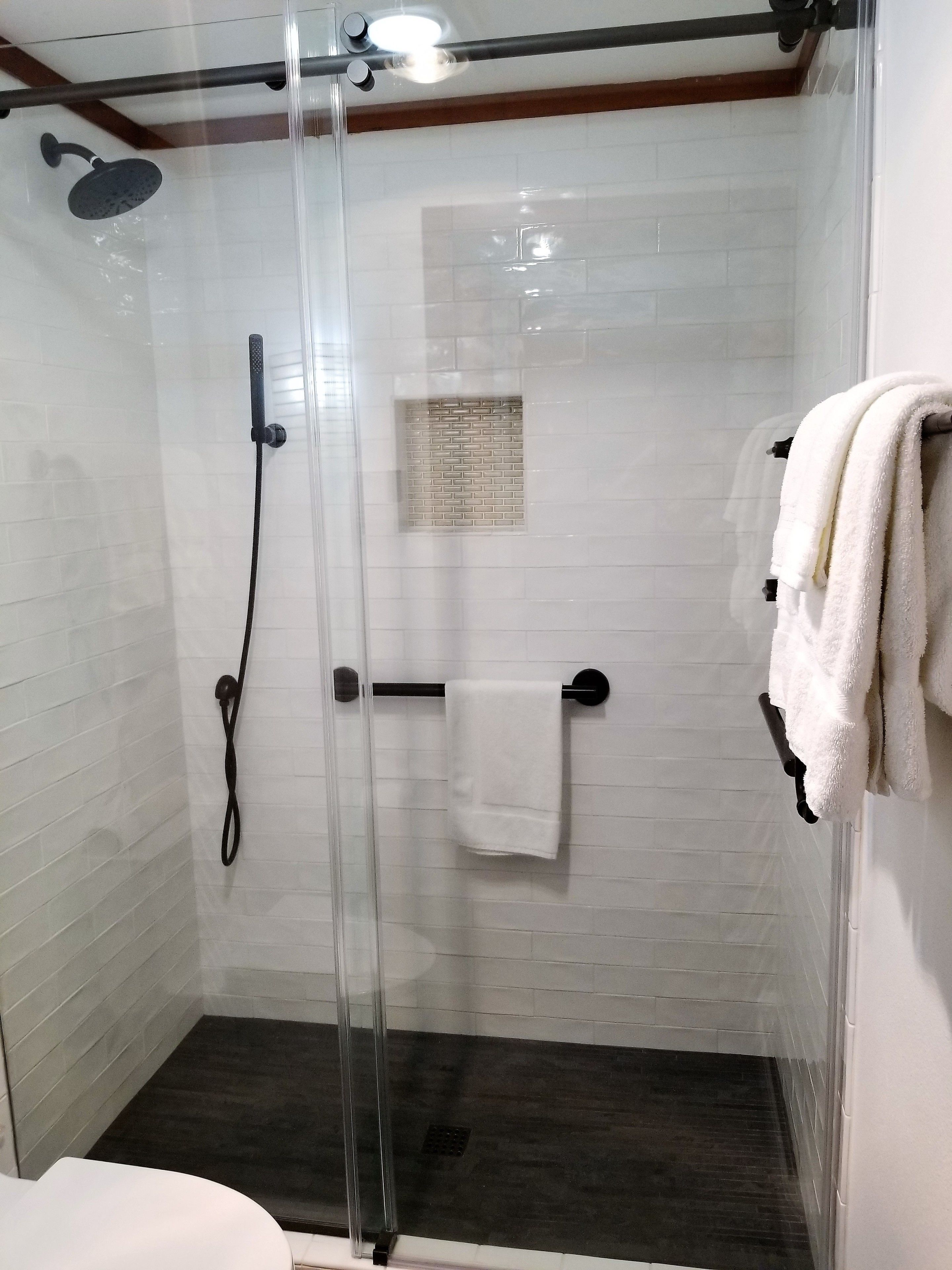 Een douchecabine met glazen wanden, een handdouche, een handdoekhouder met een witte handdoek, en een klein raam met een bakstenen patroon.