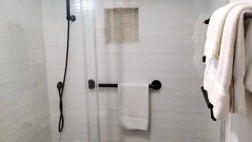 Een douchecabine met glazen wanden, een handdouche, een handdoekhouder met een witte handdoek, en een klein raam met een bakstenen patroon.