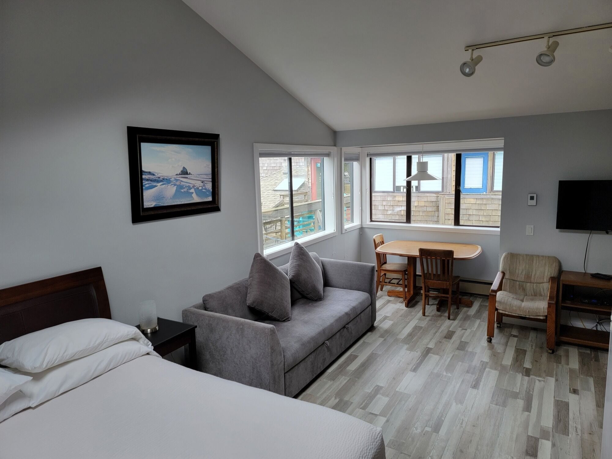 Une chambre d’hôtel avec un lit, un canapé gris, une table à manger en bois avec des chaises, une télévision et de grandes fenêtres donnant sur l’extérieur.