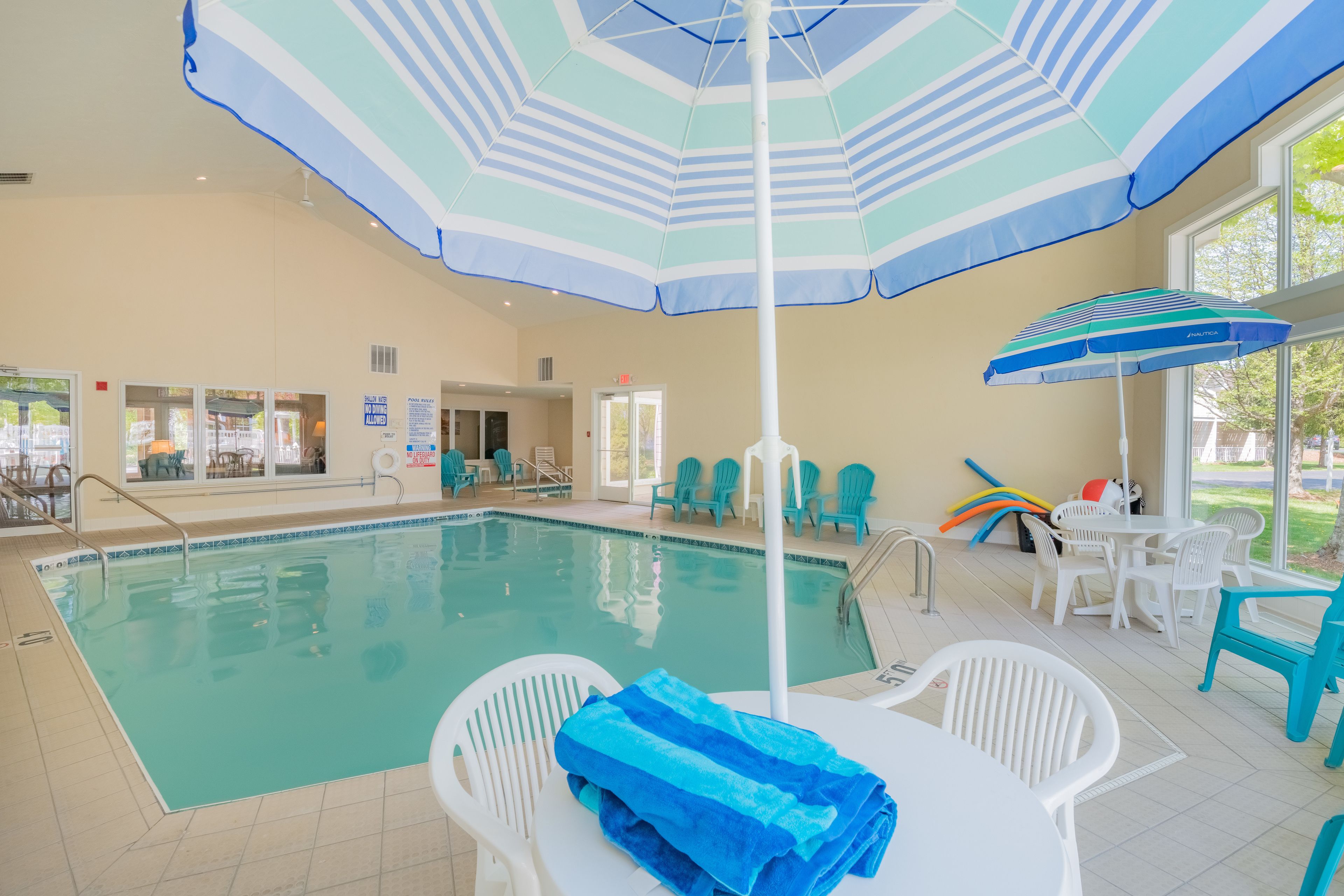 Une piscine intérieure avec un parasol bleu et blanc à rayures, des chaises blanches et une table sur laquelle est posée une serviette bleue.