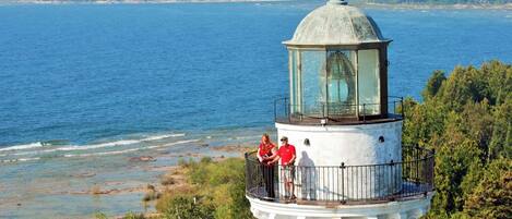 Un faro con cerramiento de cristal, rodeado de vegetación y con vistas a una masa de agua.