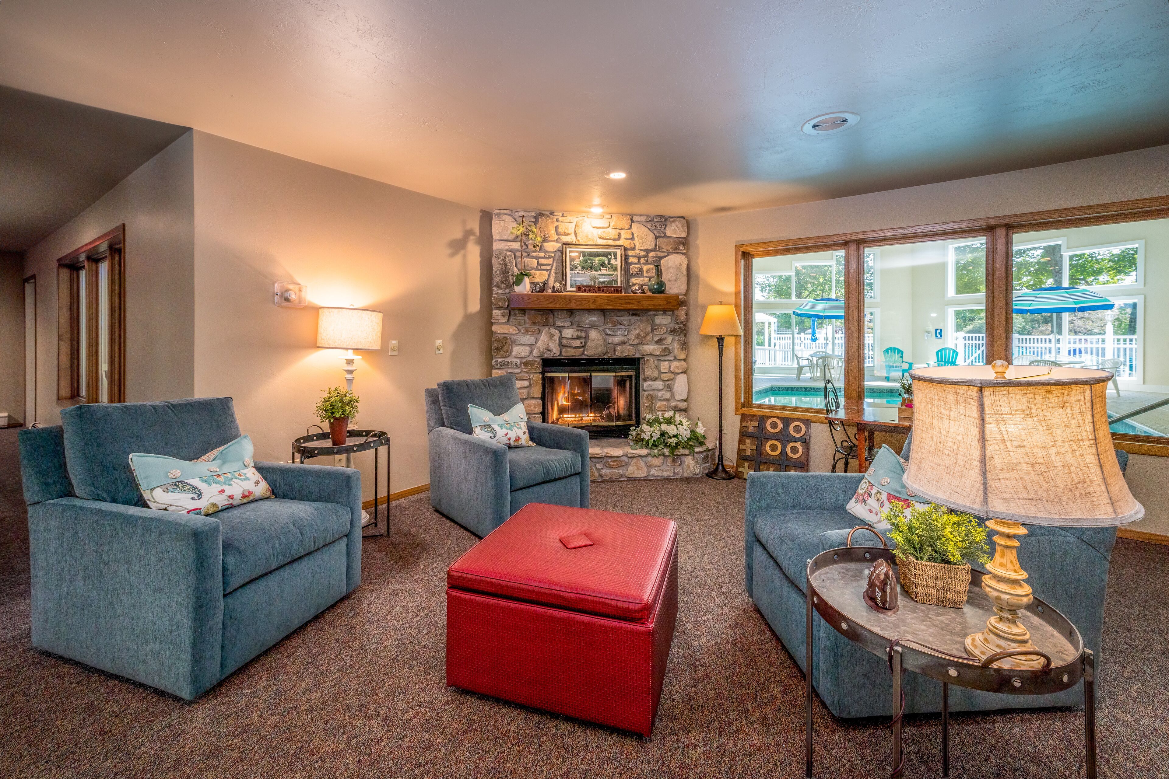 A cozy living room with two blue armchairs, a red ottoman, a stone fireplace, and a large window with a view of a deck.